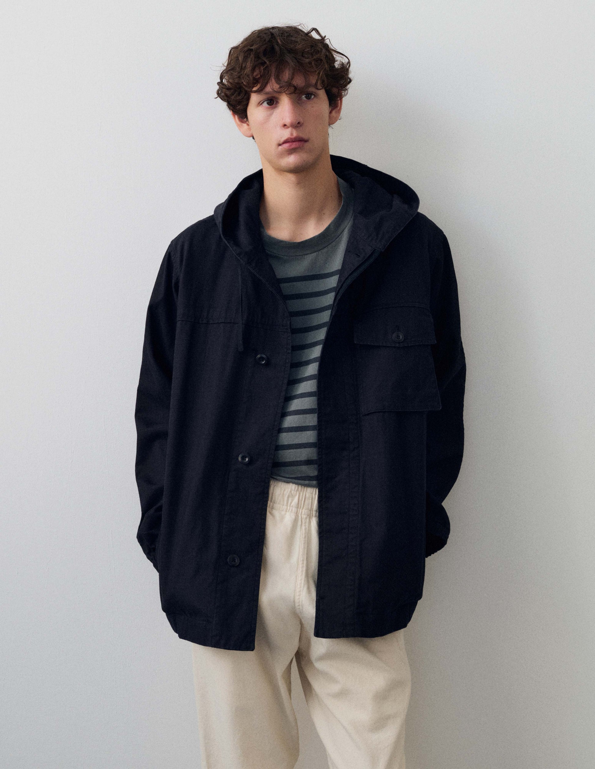 Full body image, back view of model wearing indigo blue twill hooded jacket, with sleeve patches. Styled with grey and navy striped t shirt and white trousers.