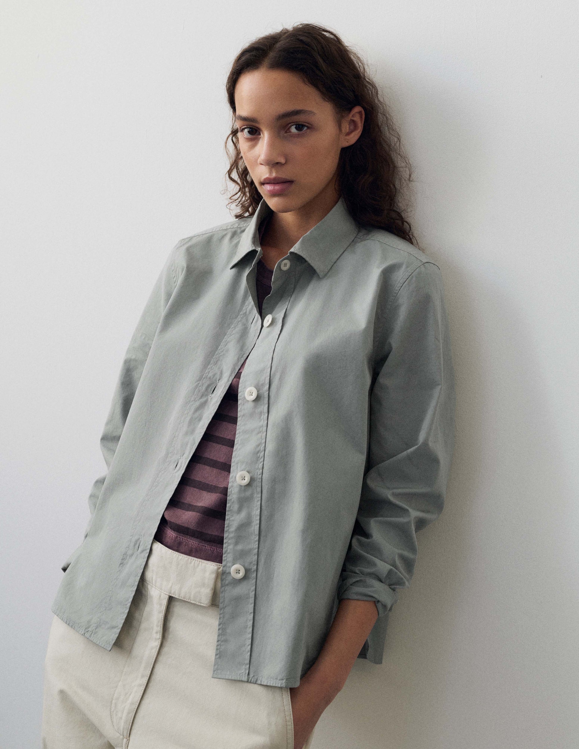 Cropped body image, front view of model wearing grey regular cut shirt with stitch through raised placket with corozo buttons on placket and cuffs. Styled with purple and brown striped t shirt and white trousers.