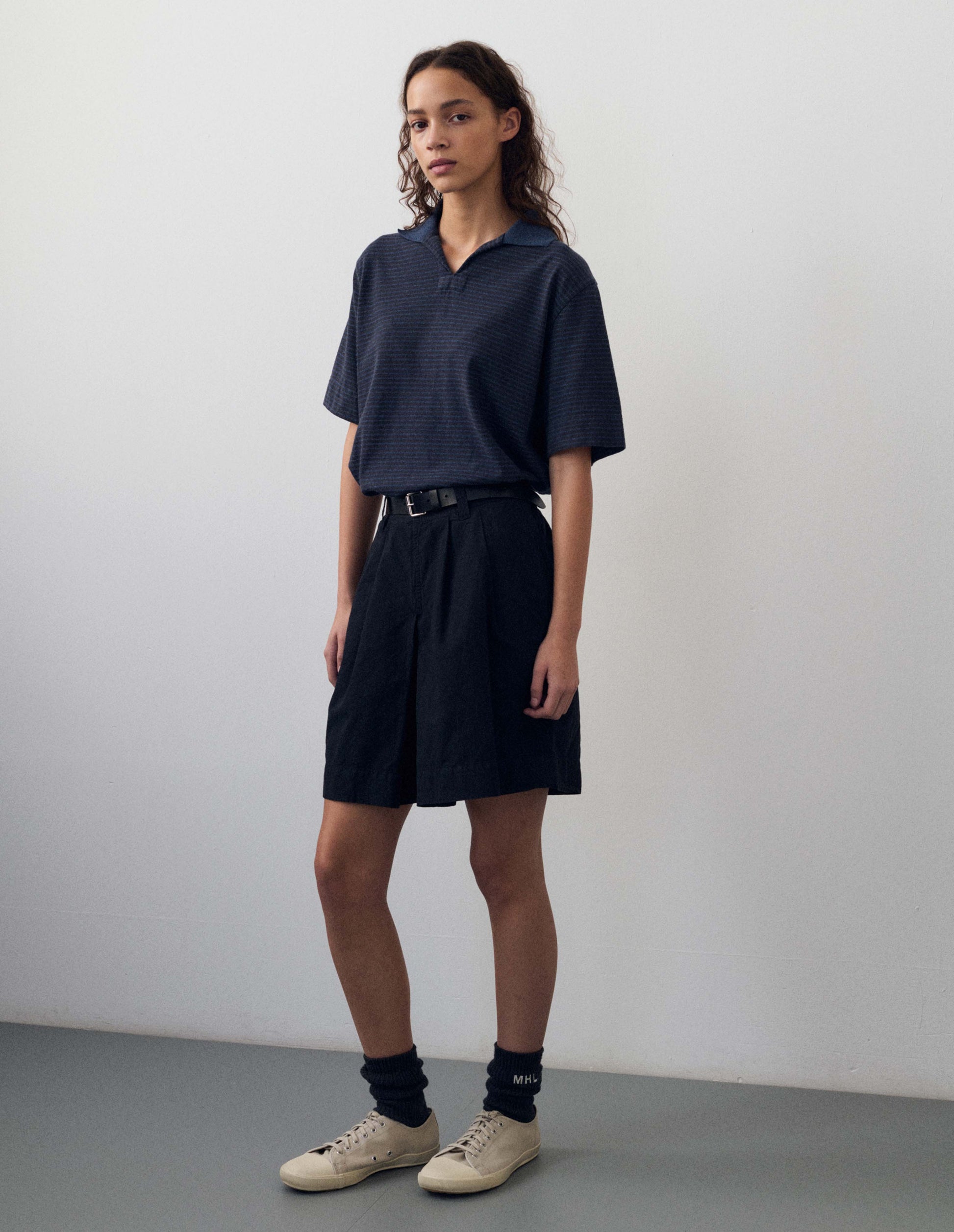 Full body image, front view of model wearing dark blue and light blue striped polo t-shirt, with open neck contrast melange rib collar and side split hem. Styled with dark navy blue shorts, black socks and white canvas trainers.