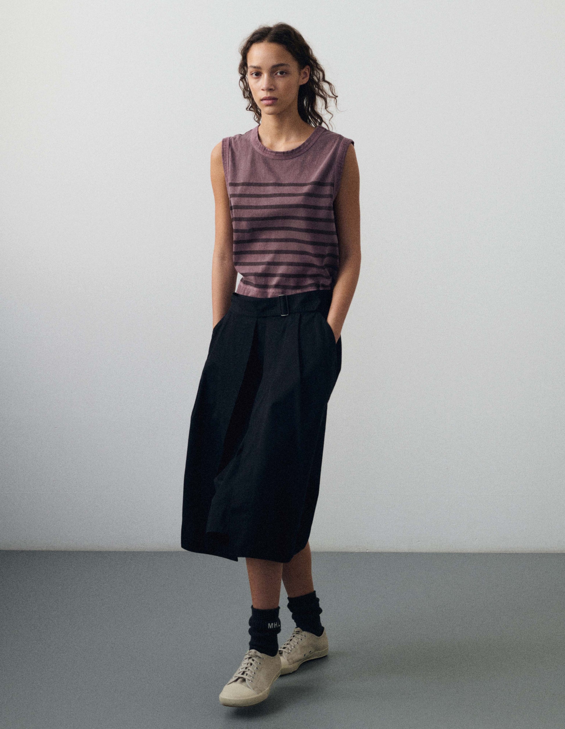 Full body image, front view of model wearing purple cotton linen tank top with brown stripes, contrast ribbing on neckline and armholes. Styled tucked into black skirt, with black socks and beige canvas trainers.