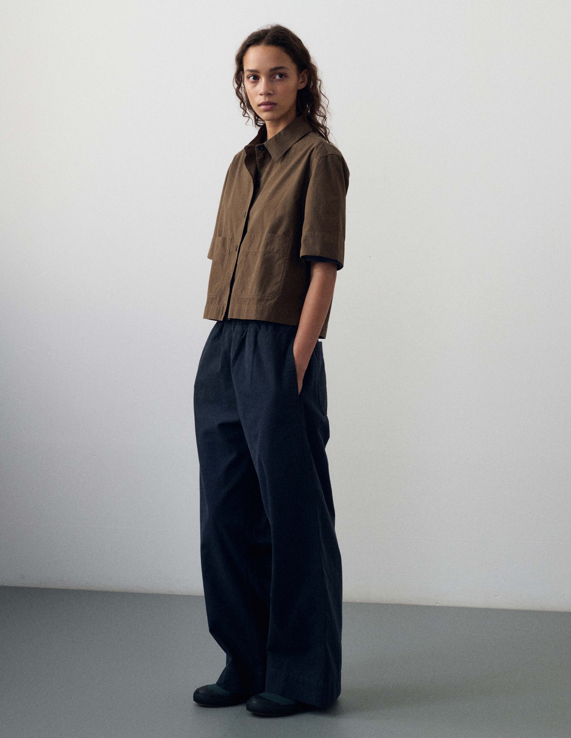 Full body image, front view of model wearing dark navy blue joggers with elasticated waistband and side seam pockets. Styled with brown short sleeve shirt and dark blue canvas shoes with black gum soles. 