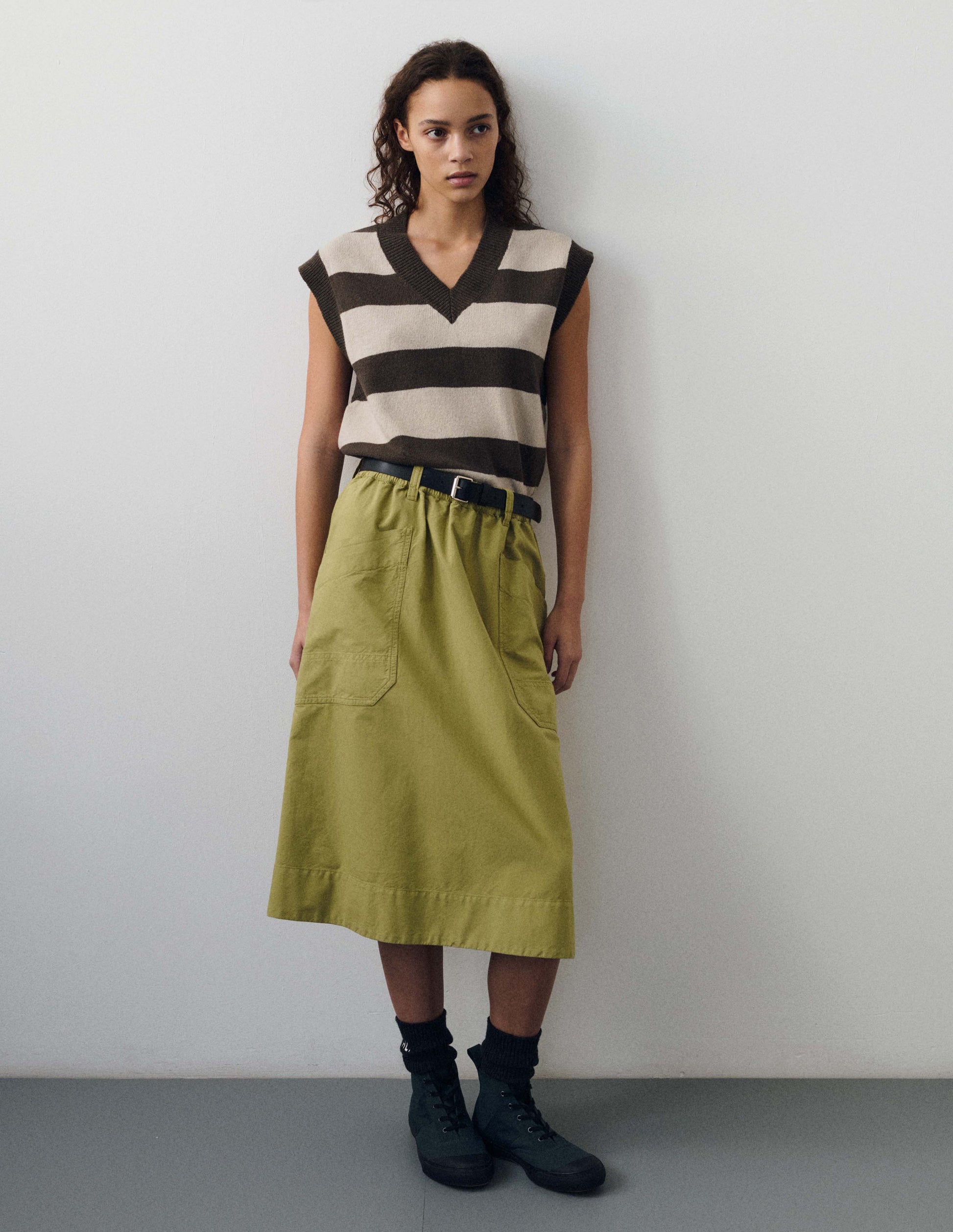 Full body image, front view of model wearing brown and off white chunky striped sleeveless knitted jumper, with thick brown rib on v neck, armholes and hem. Styled with green sjirt, black leather belt, black socks and dark blue high top trainers with black gum sole.