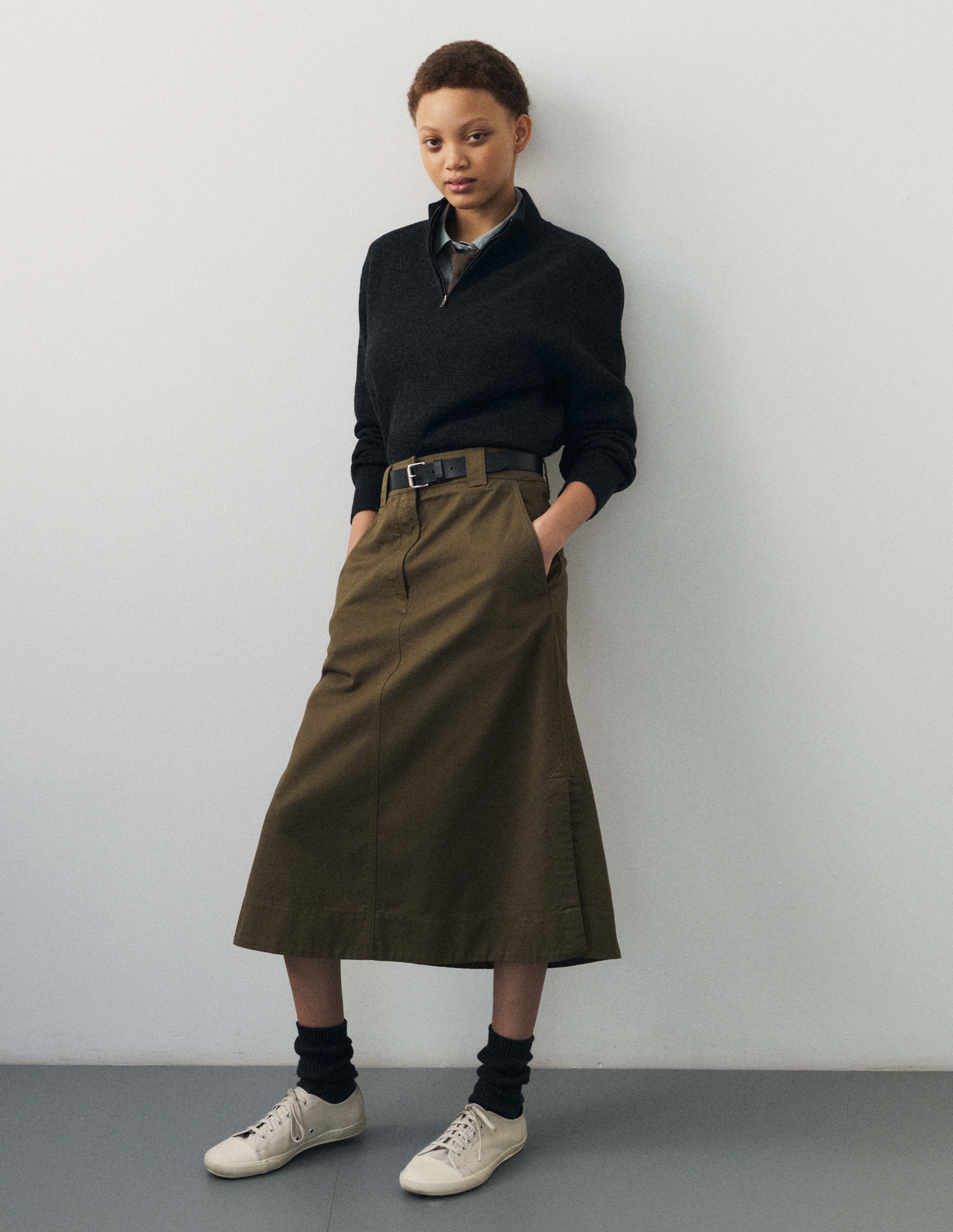 Full body image, front view of model wearing dark grey wool cotton half zip sweater with contrast rib on neck and cuffs, and contrast white line around ribbed hem. Styled with bronwish green long skirt, black socks and white canvas trainers. 