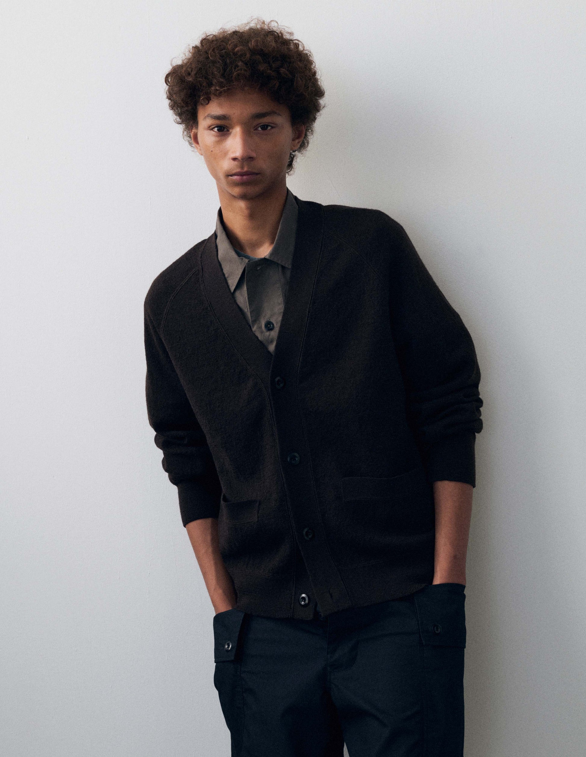 Full body image, front view of model wearing dark grey v neck wool cardigan, with concealed pockets, corozo button closure, raglan sleeves and contrast rib on cuffs and hem. Styled with greyish brown collared shirt and dark navy blue cargo trousers.