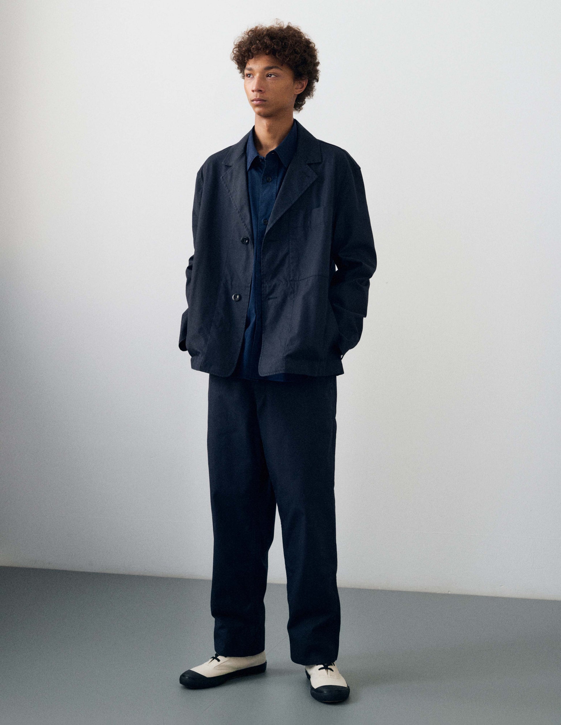 Full body image, front view of model wearing dark navy blue cotton blazer, with horn button fastening lapel, large patch chest pocket and side pockets. Styled with dark navy blue shirt, dark navy blue trousers and cream canvas trainers with black gum soles.