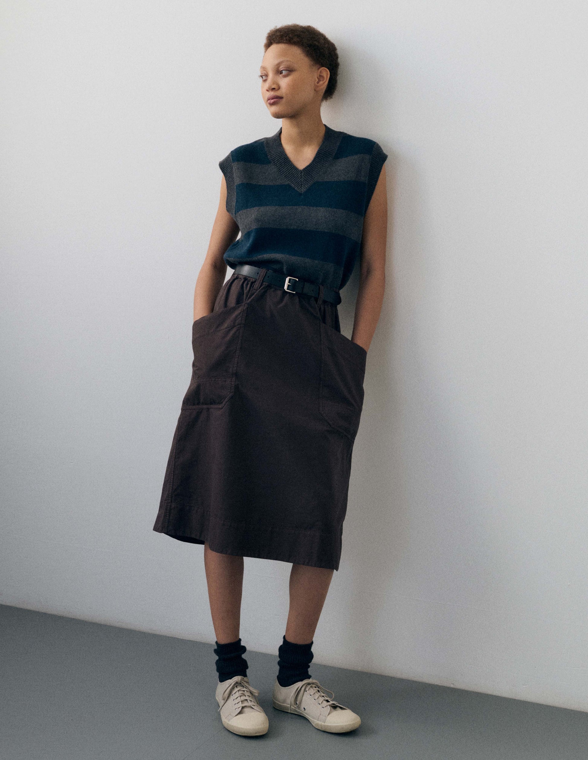 Full body image, front view of model wearing grey and dark navy striped v neck slipover, with contrast rib v neck, armholes and hem. Styled with purplish brown skirt, black leather belt, black sock and white canvas trainers.