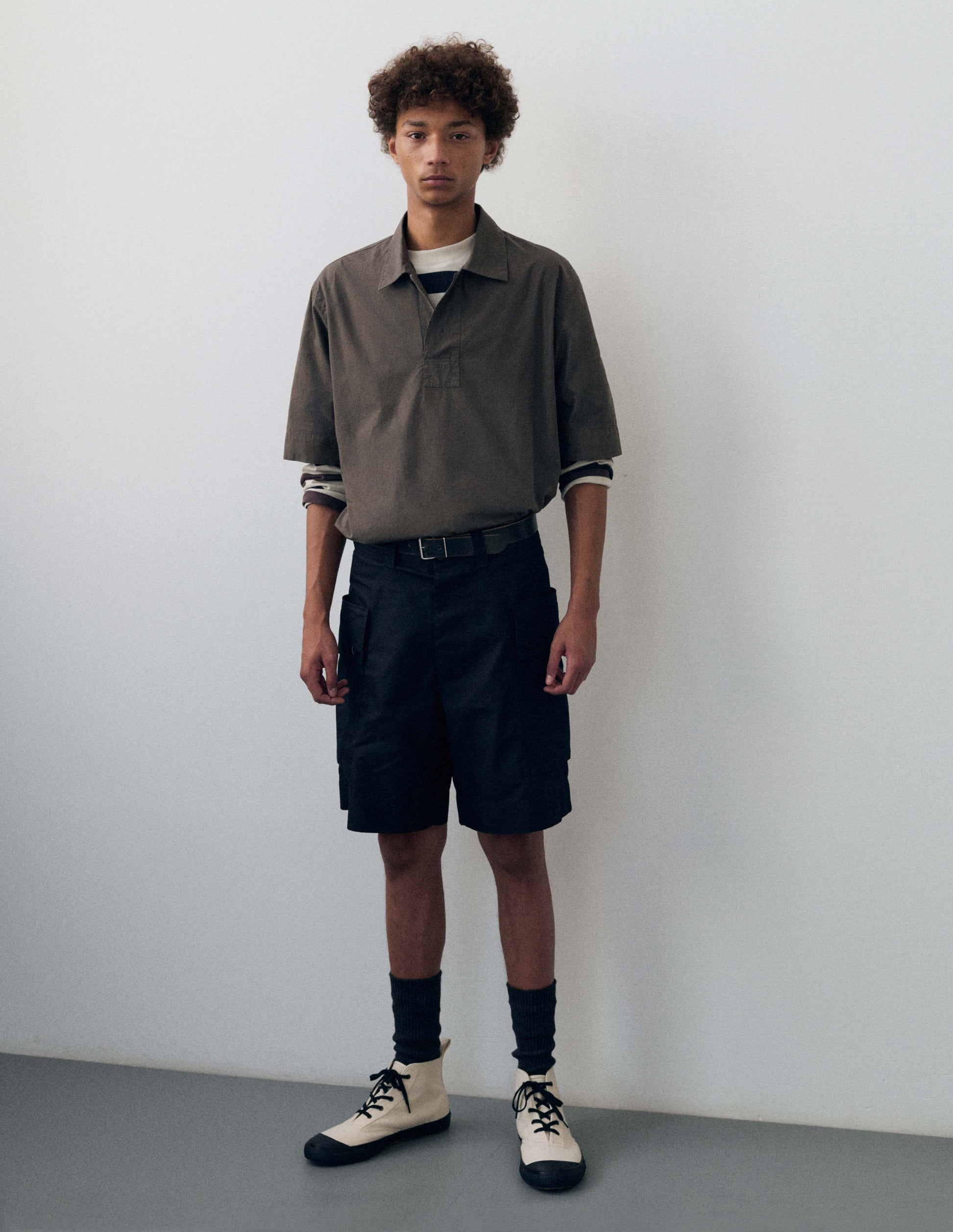 Full body image, front view of model wearing black cotton shorts with cargo pockets, belt loops and welt back pocket.  Styled with wite and brown striped long sleeve t-shirt and brown short sleeve collared shirt tucked in, with black socks and white high top canvas trainers. 