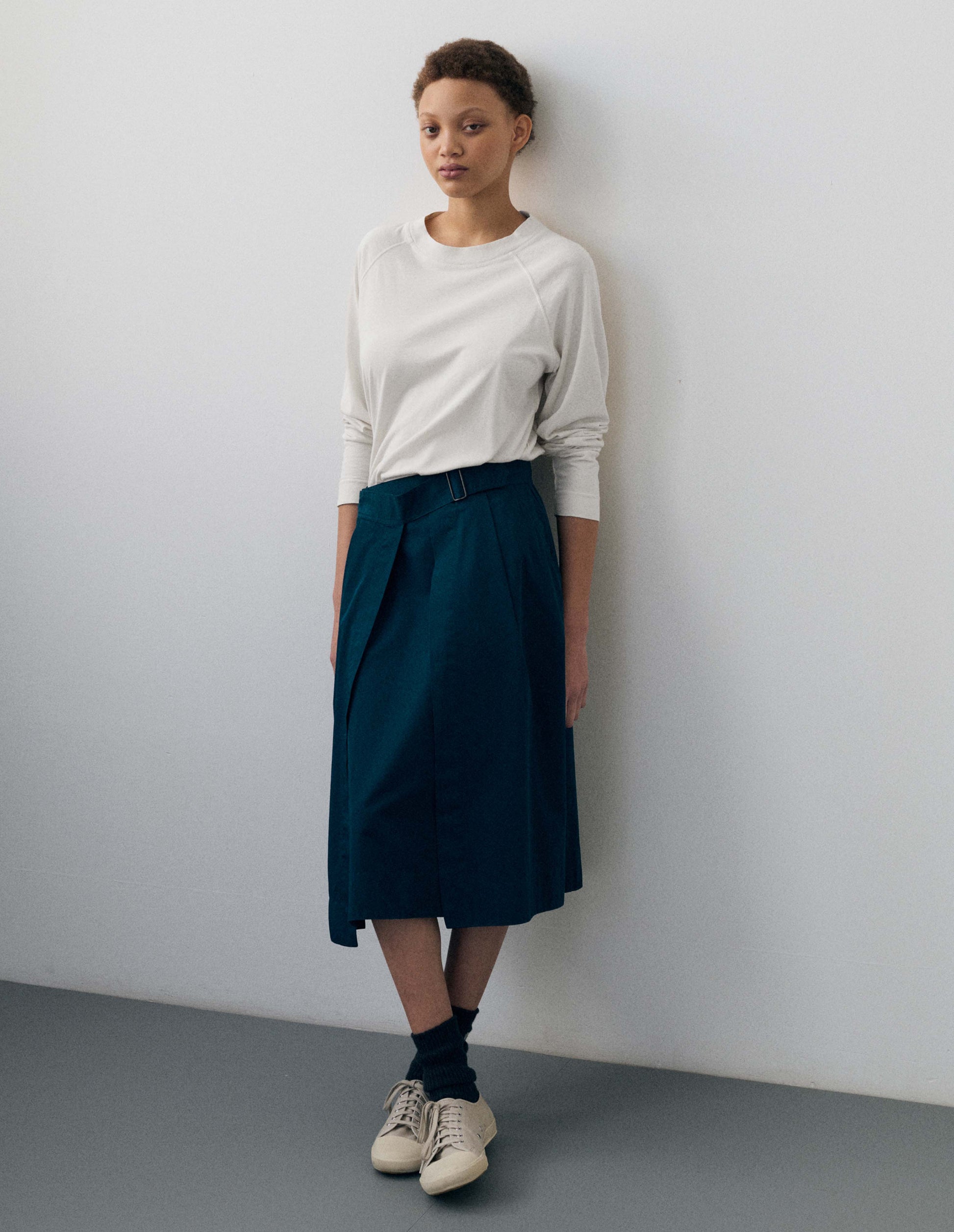 Full body image, front view of model wearing greenish blue cotton twill skirt, with wide crossover waistband, button fastening slide adjuster and slant pockets. Styled with white long sleeve t shirt, black socks and beige canvas trainers.