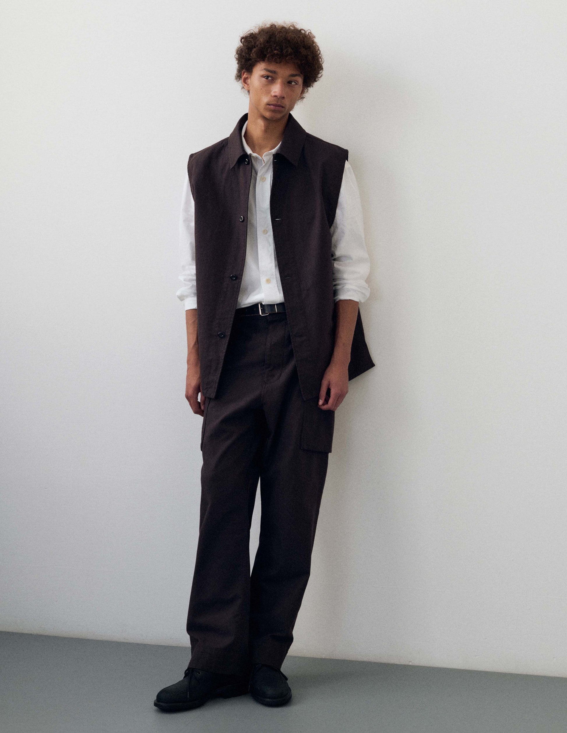 Full body image, front view of model wearing dark brown cotton drill sleeveless overshirt with corozo button fastening and vent split hem. Styled with white shirt, black trousers, black leather belt and black leather shoes. 