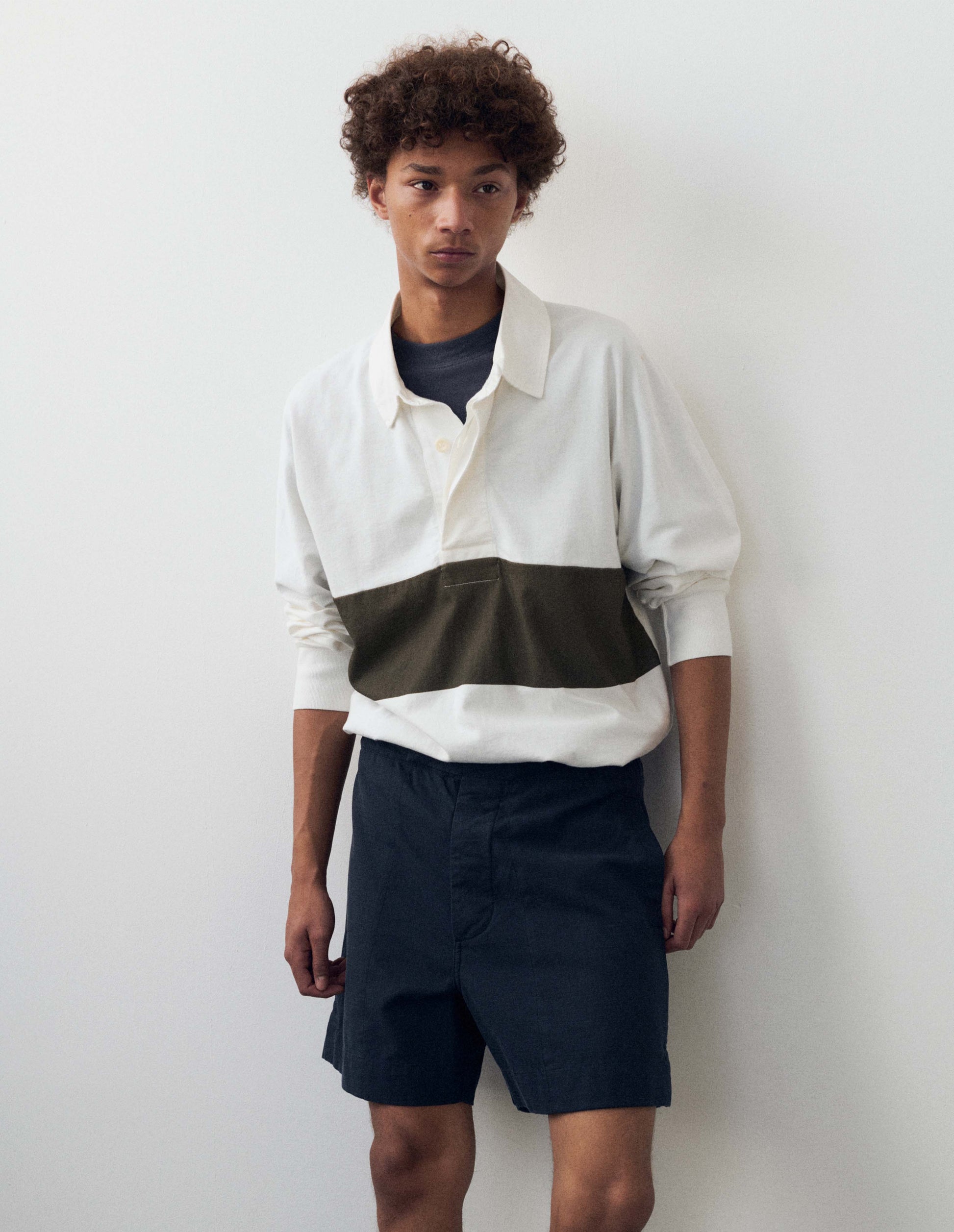 Cropped body image, front view of model wearing off white rugby long sleeve polo shirt with dark green panel across chest, twill collar, wide fly front placket and rib cuffs. Styled with dark navy blue t shirt and dark navy blue shorts. 