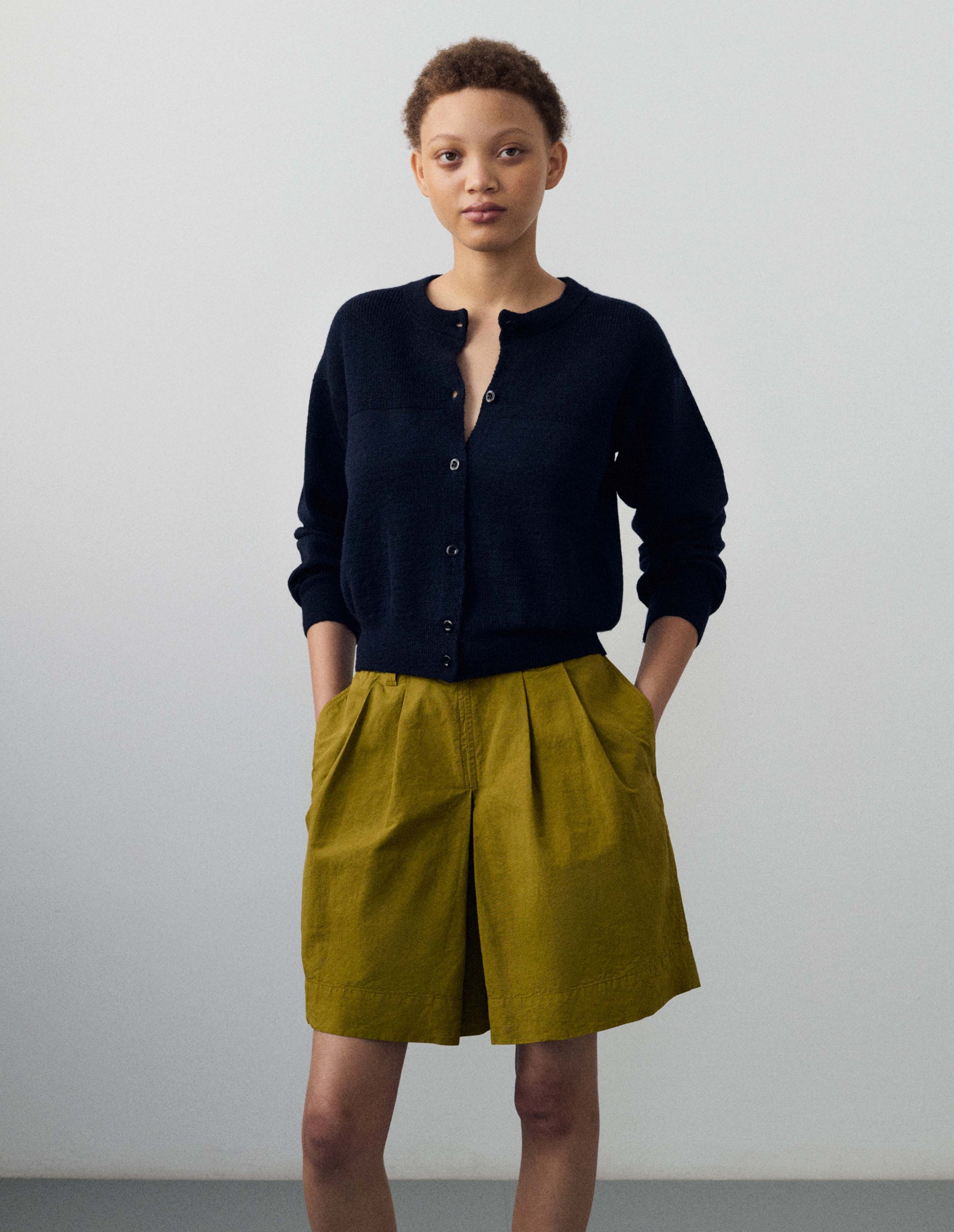 Cropped body image, front view of model wearing dark navy blue wool textured cardigan, with contrast panel across chest, rib detailing on neck, cuffs and hem and corozo button closure. Styled with green shorts. 