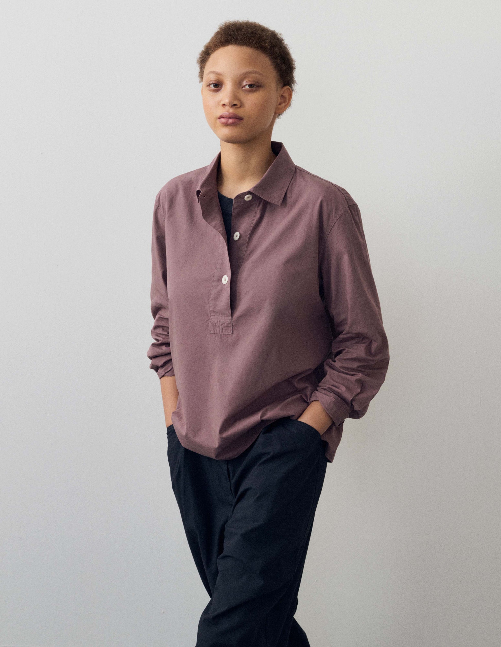 Cropped body image, front view of model wearing purpleish pink cotton poplin swing shirt, with half raised placket and corozo button fastening. Styled with dark navy blue trousers.