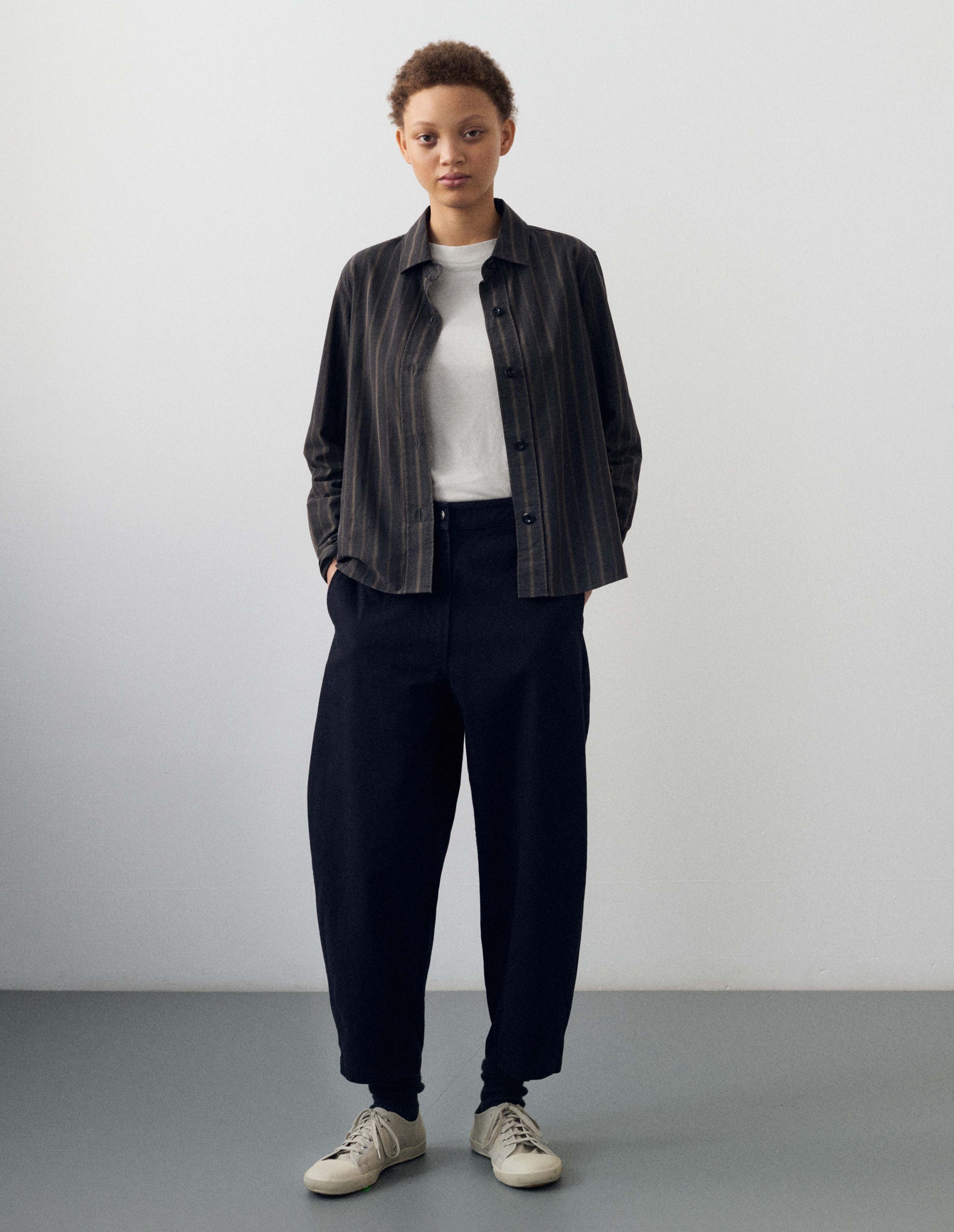 Full body image, front view of model wearing indigo blue barrel leg trouser, with slant side pockets and silver metal closure. Styled with white t shirt, black and brown pinstripe long sleeve collared shirt, black socks and white canvas trainers.