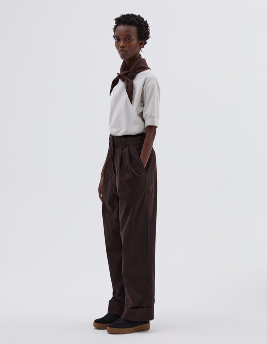 Full body image, side view of model wearing dark brown pleated trousers with zip fly and double hook and bar fastening, slant side pockets and turn up hems. Styled with white t-shirt tucked in, brown knitted scout scarf and black suede trainers.