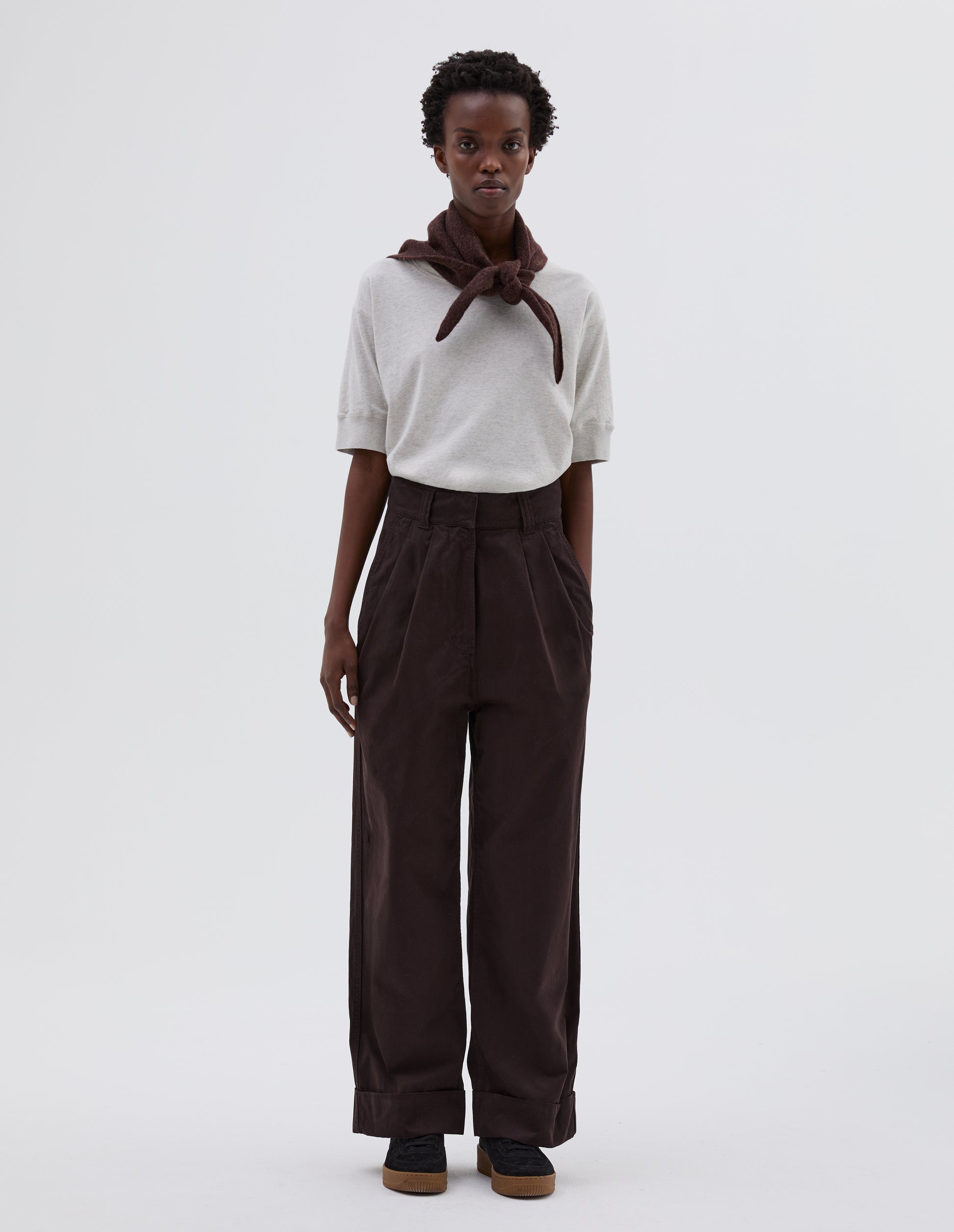 Full body image, front view of model wearing dark brown pleated trousers with zip fly and double hook and bar fastening, slant side pockets and turn up hems. Styled with white t-shirt tucked in, brown knitted scout scarf and black suede trainers.