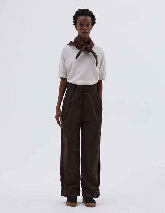 Full body image, front view of model wearing dark brown pleated trousers with zip fly and double hook and bar fastening, slant side pockets and turn up hems. Styled with white t-shirt tucked in, brown knitted scout scarf and black suede trainers.