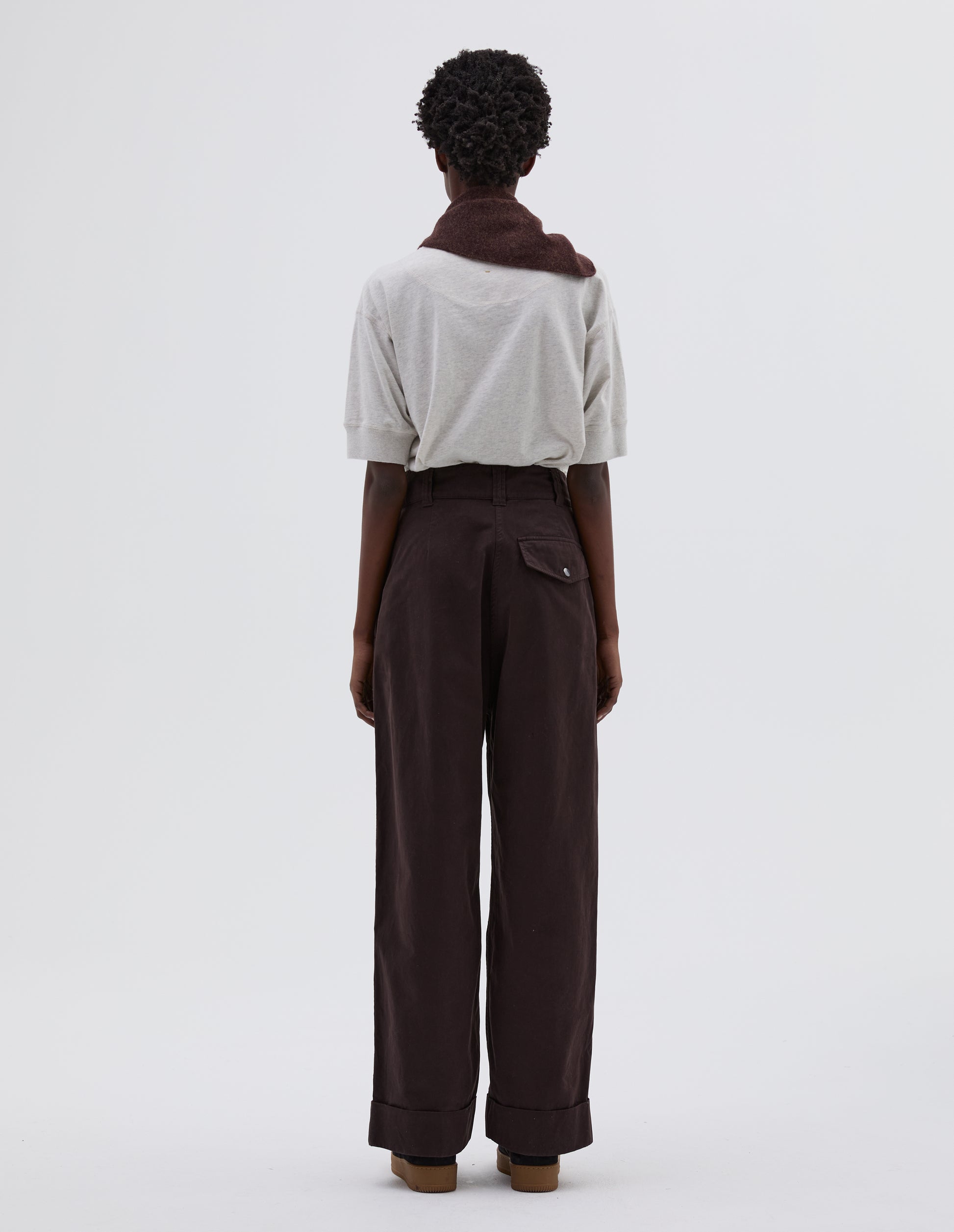 Full body image, back view of model wearing dark brown pleated trousers with popper fastening back flap pocket and belt loops. Styled with white t-shirt tucked in, brown knitted scout scarf and black suede trainers.