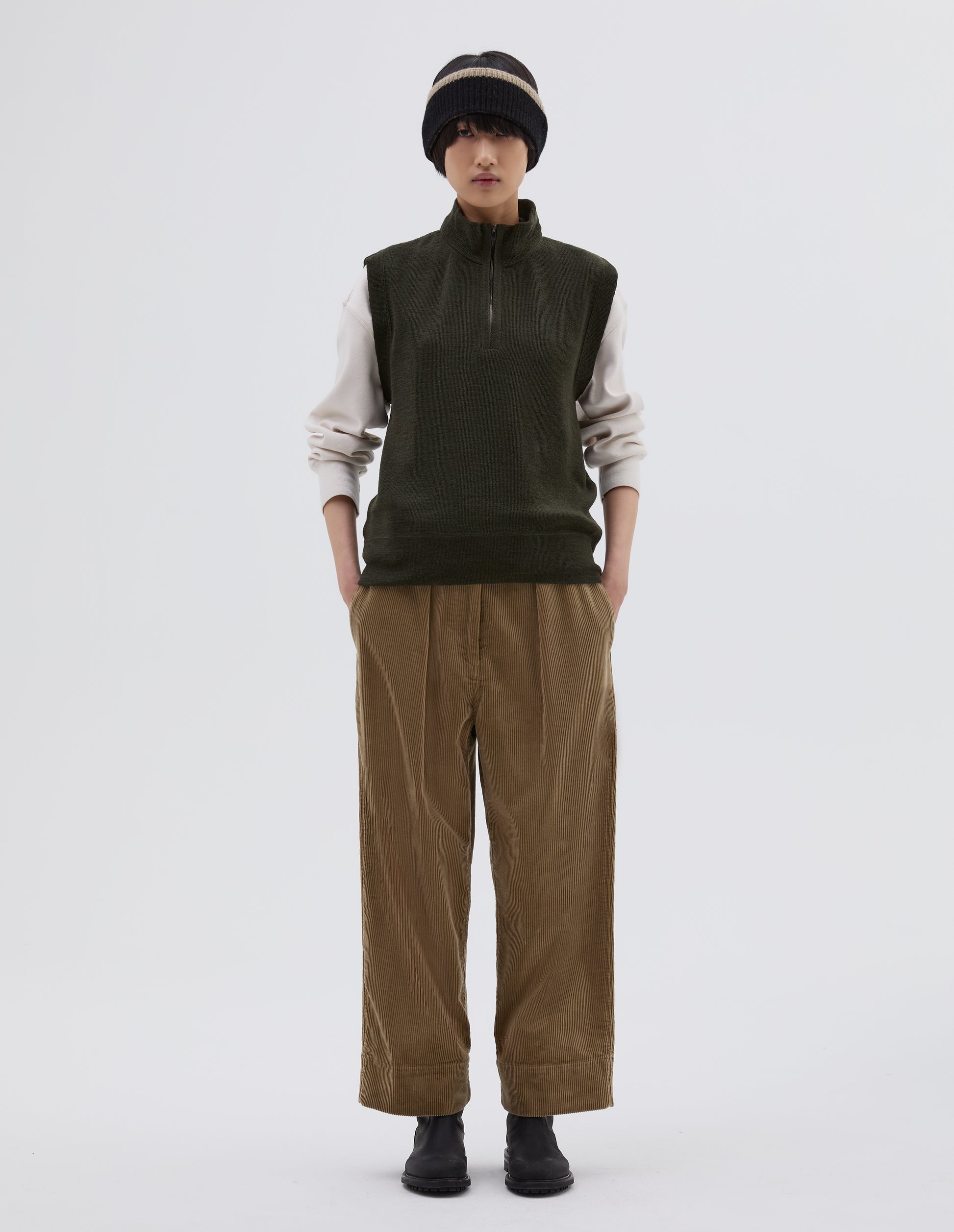 Full body image, front view of model wearing dark green wool slipover, with half zip mock turtleneck and flatlock stitch detailing on seams. Styled over cream sweatshirt, with black beanie hat, beige corduroy trousers and black leather boots.
