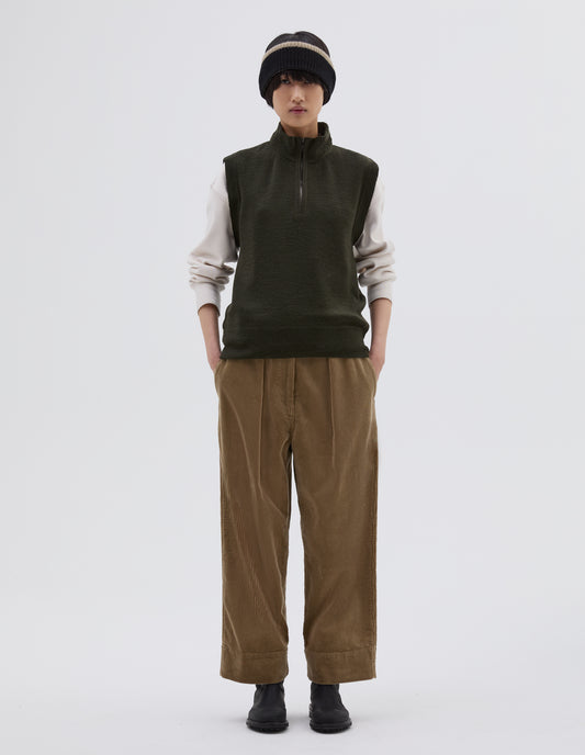Full body image, front view of model wearing dark green wool slipover, with half zip mock turtleneck and flatlock stitch detailing on seams. Styled over cream sweatshirt, with black beanie hat, beige corduroy trousers and black leather boots.