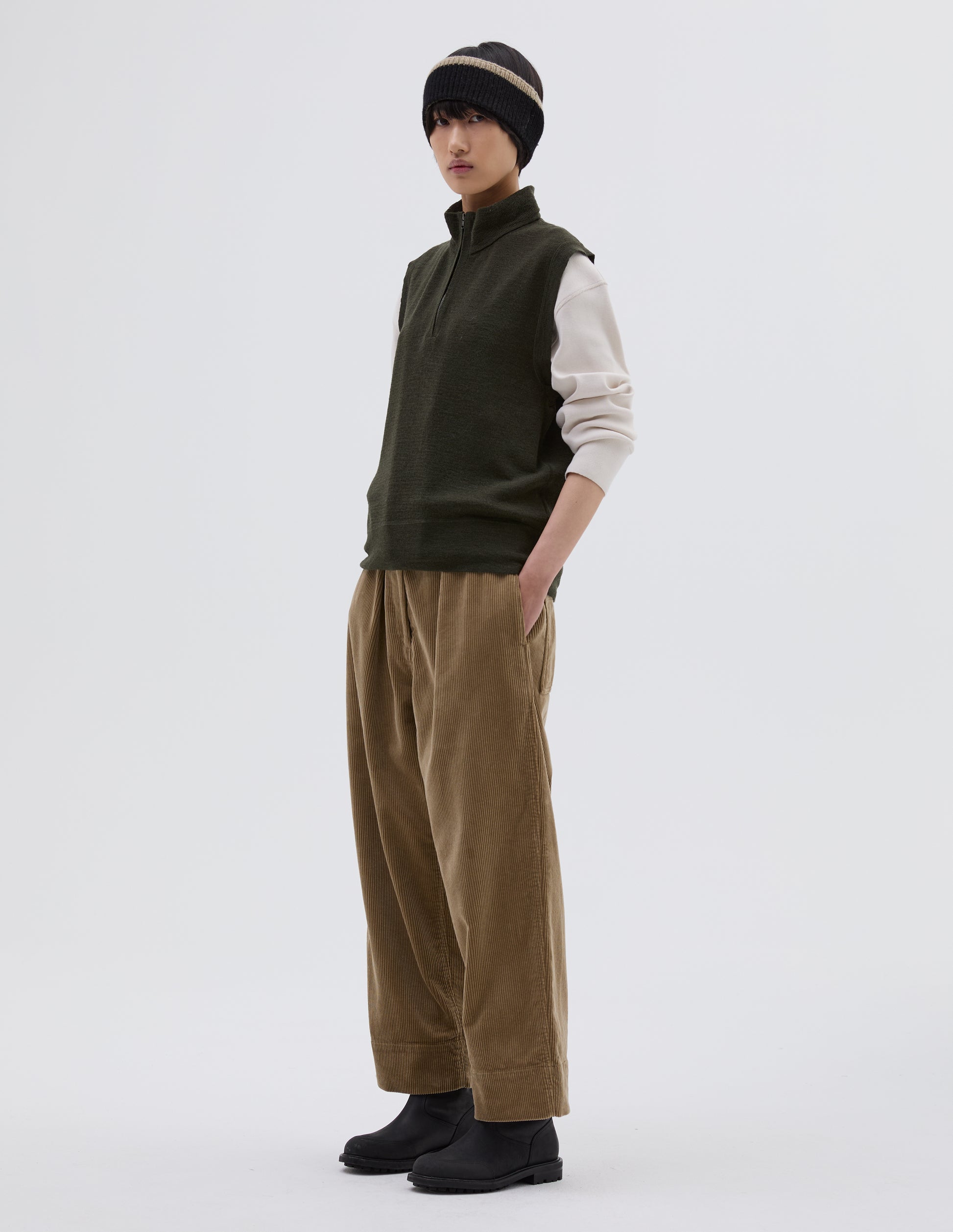 Full body image, side view of model wearing dark green wool slipover, with half zip mock turtleneck and flatlock stitch detailing on seams. Styled over cream sweatshirt, with black beanie hat, beige corduroy trousers and black leather boots.