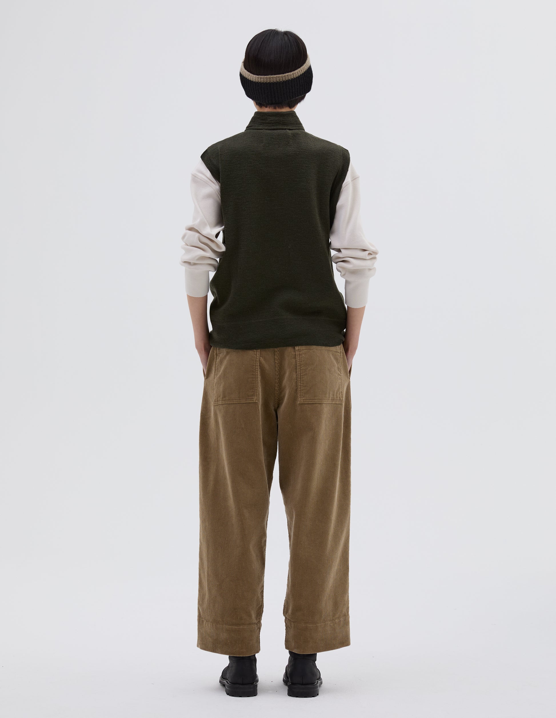 Full body image, back view of model wearing dark green wool slipover, with half zip mock turtleneck and flatlock stitch detailing on seams. Styled over cream sweatshirt, with black beanie hat, beige corduroy trousers and black leather boots.